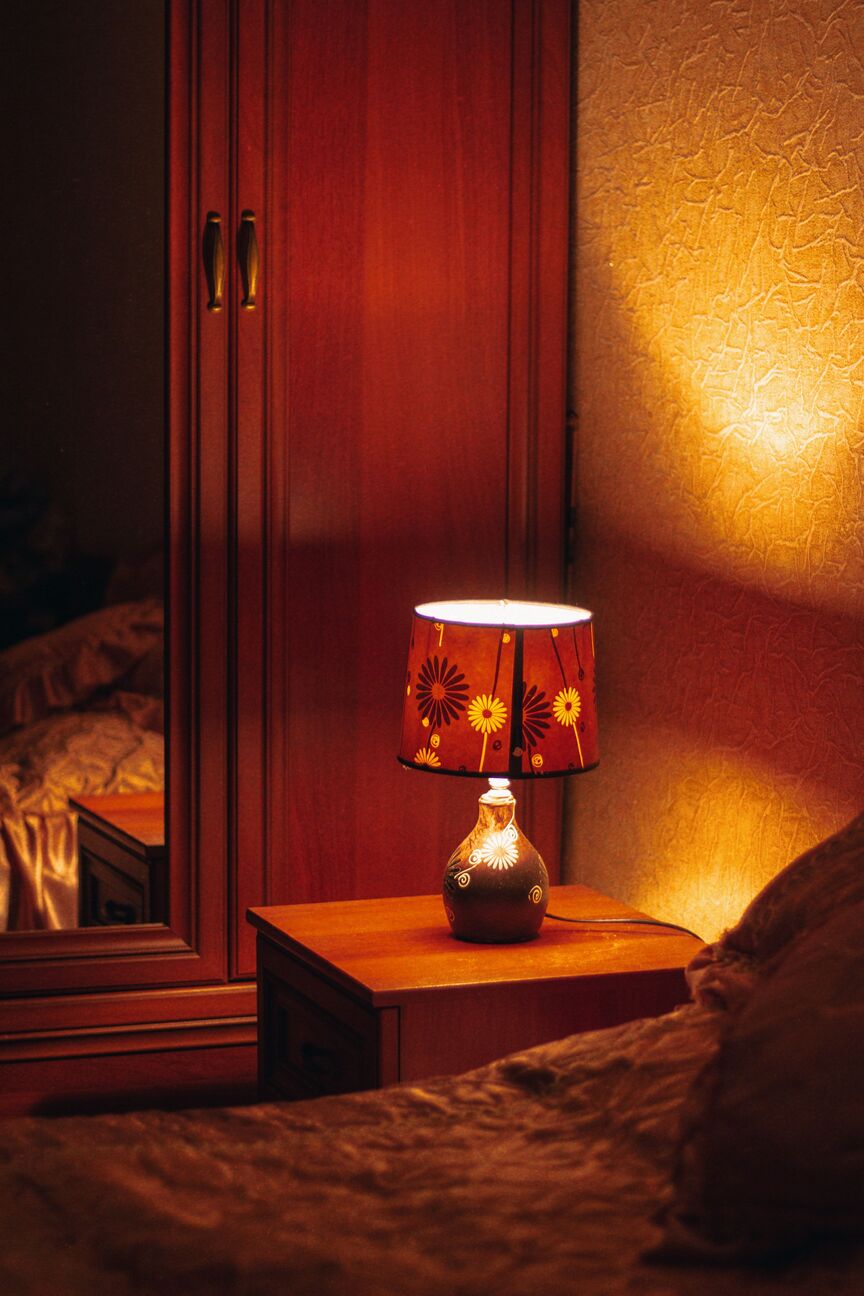 Warm bedside lamp glowing on a wooden nightstand beside a bed in a dim evening bedroom