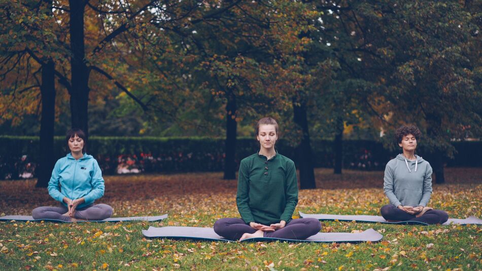 Three people sitting calmly on mats in an autumn park, practising slow breathing