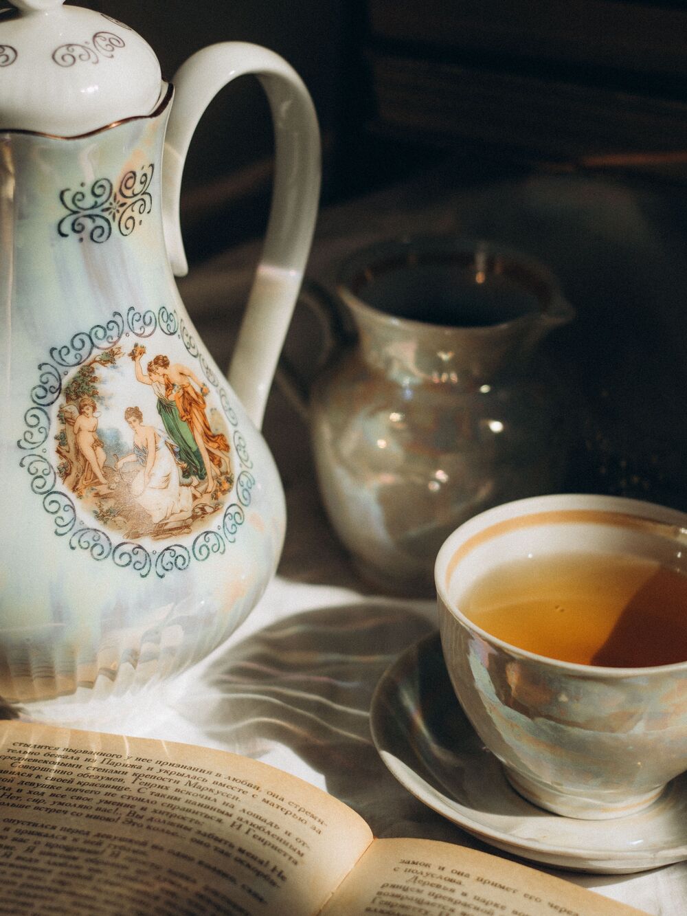 Porcelain teapot and a cup of tea beside an open book on a sunlit table